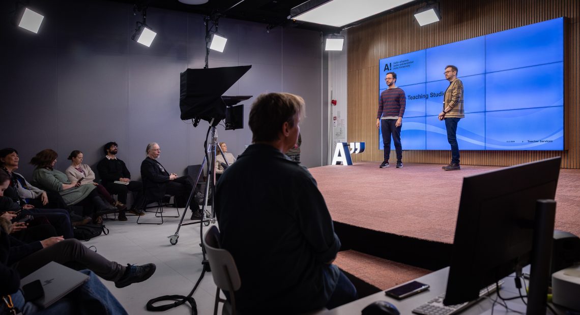 Two people stand on the stage in front of a large, wall-mounted TV screen displaying a blue slide with the text "Testing the Teaching Studio." The stage is surrounded by a group of workshop-participating teacher colleagues.